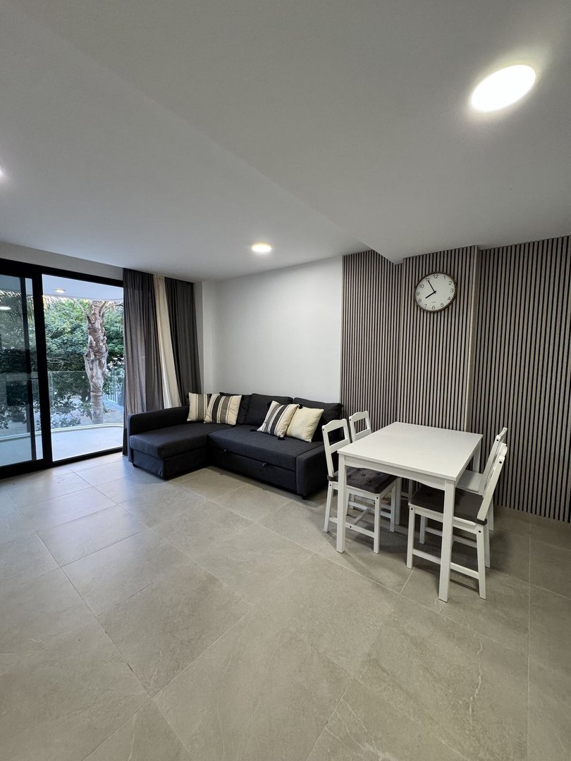 Modern living room with a dark sectional sofa, white dining table, and a clock on a slatted wall. Large window offers a view of greenery.