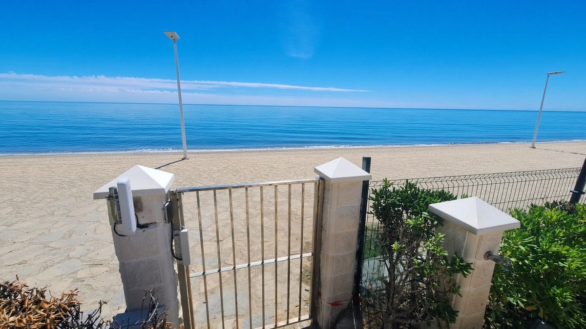 Metal gate opens to a sandy beach and calm blue ocean under a clear sky with two lampposts.
