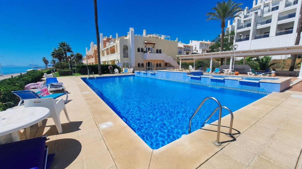 A vibrant blue communal swimming pool with loungers and palm trees, overlooking the sea under a clear sky.