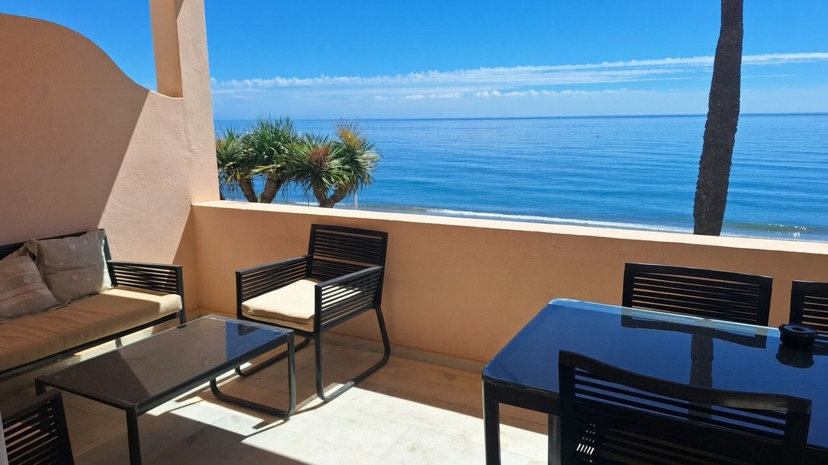 A balcony with outdoor seating and a dining table overlooks the calm blue sea on a sunny day.