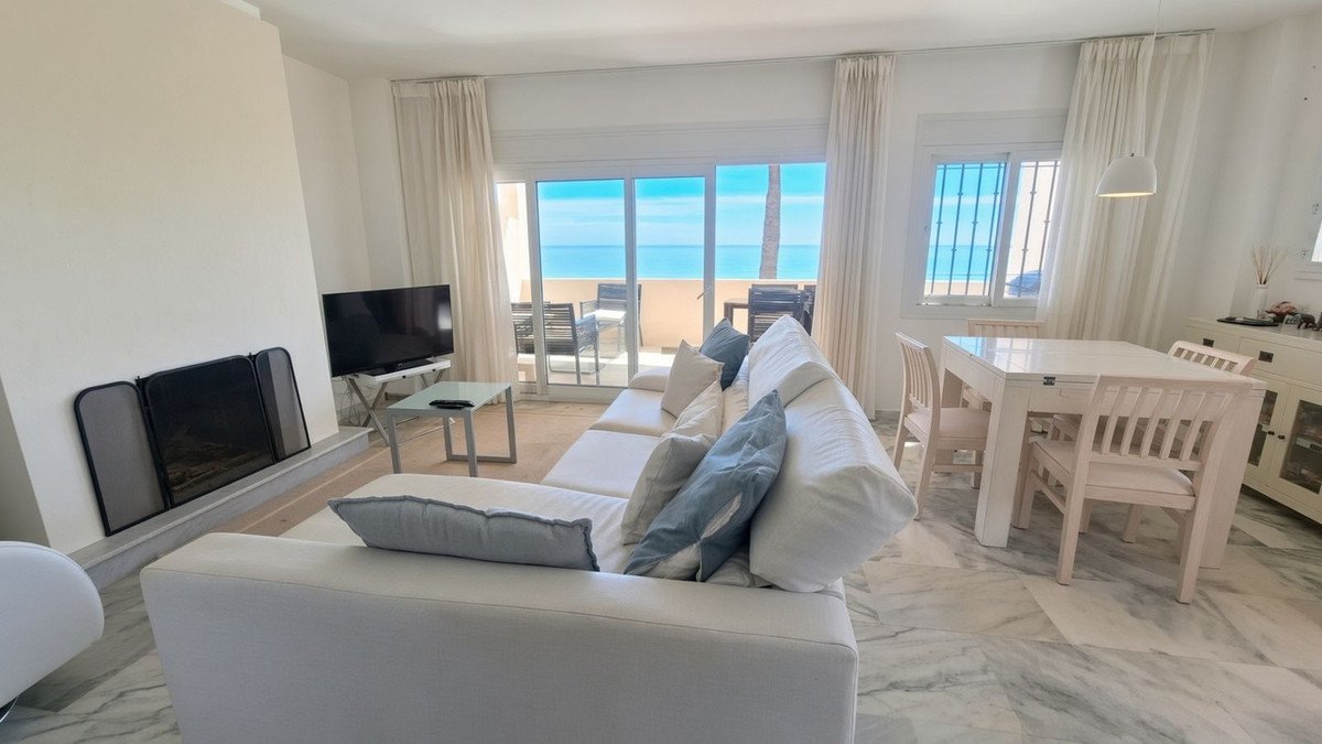 Modern living and dining area with a white sofa, fireplace, and a large balcony overlooking the sea and beach.
