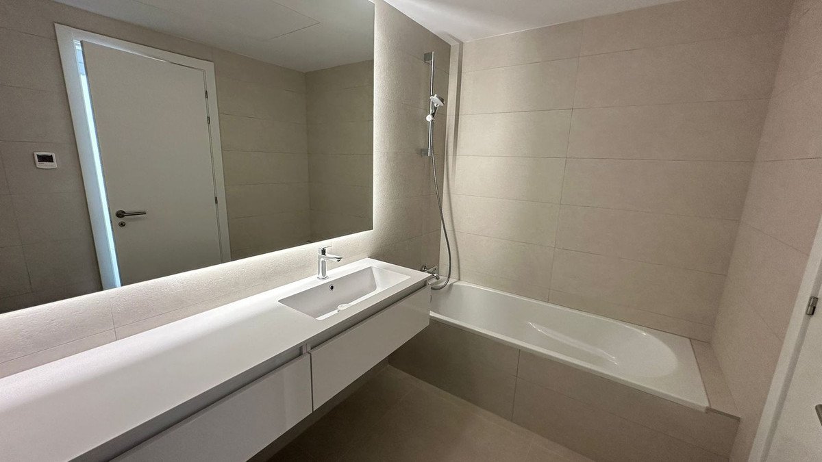 A modern bathroom featuring a long vanity with an integrated sink, a large illuminated mirror, and a bathtub with a showerhead.