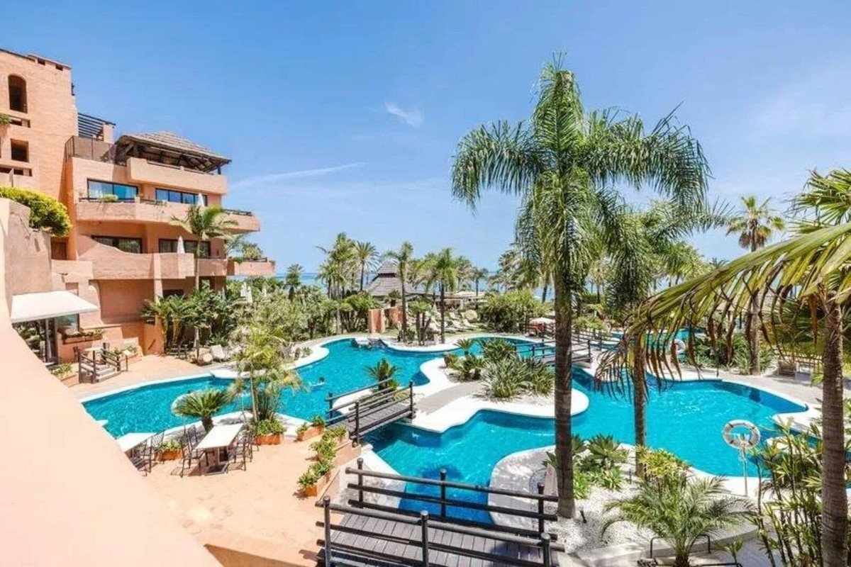 Luxurious resort with a winding pool, bridges, tropical plants, and palm trees under a clear blue sky.
