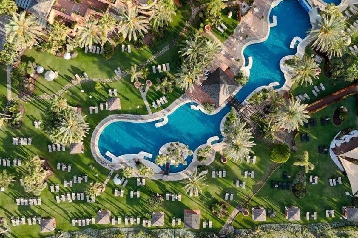 Aerial view of a resort with a large blue swimming pool, surrounded by lush green grass, palm trees, and many lounge chairs.