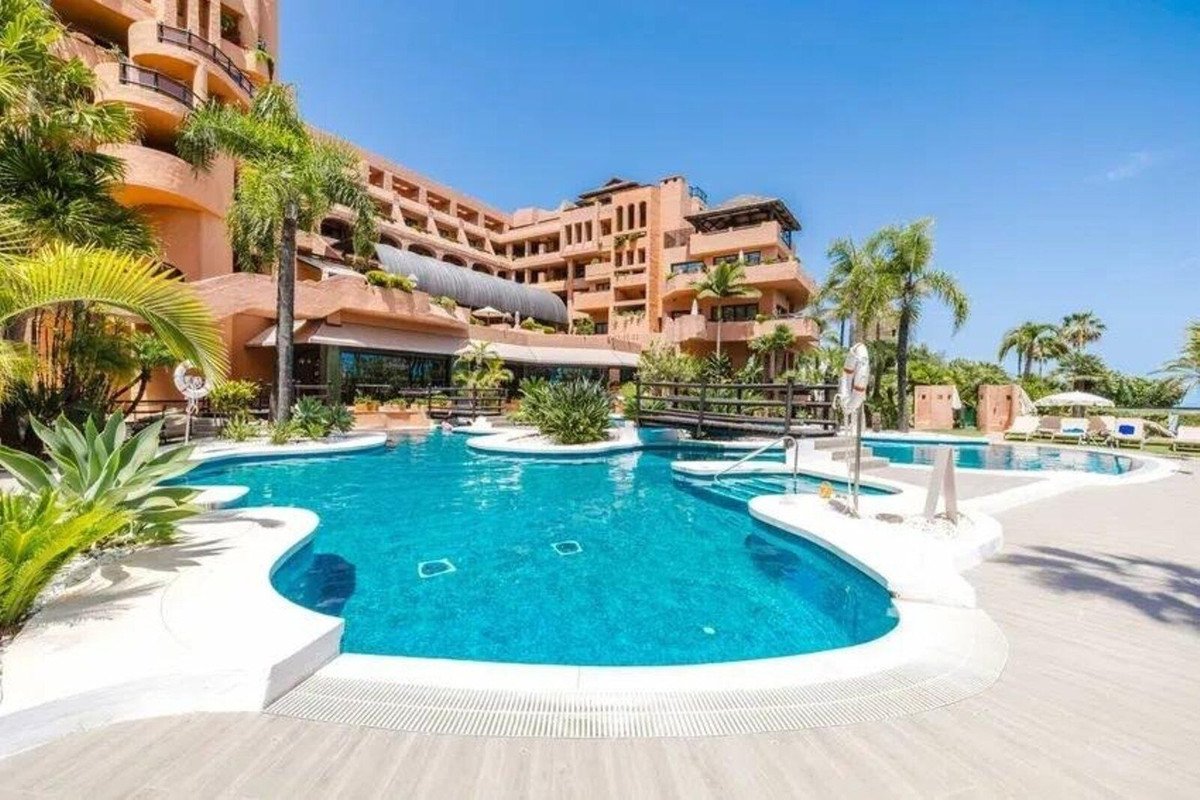 Luxurious resort complex with a large turquoise swimming pool, surrounded by palm trees and lush greenery, under a clear blue sky.