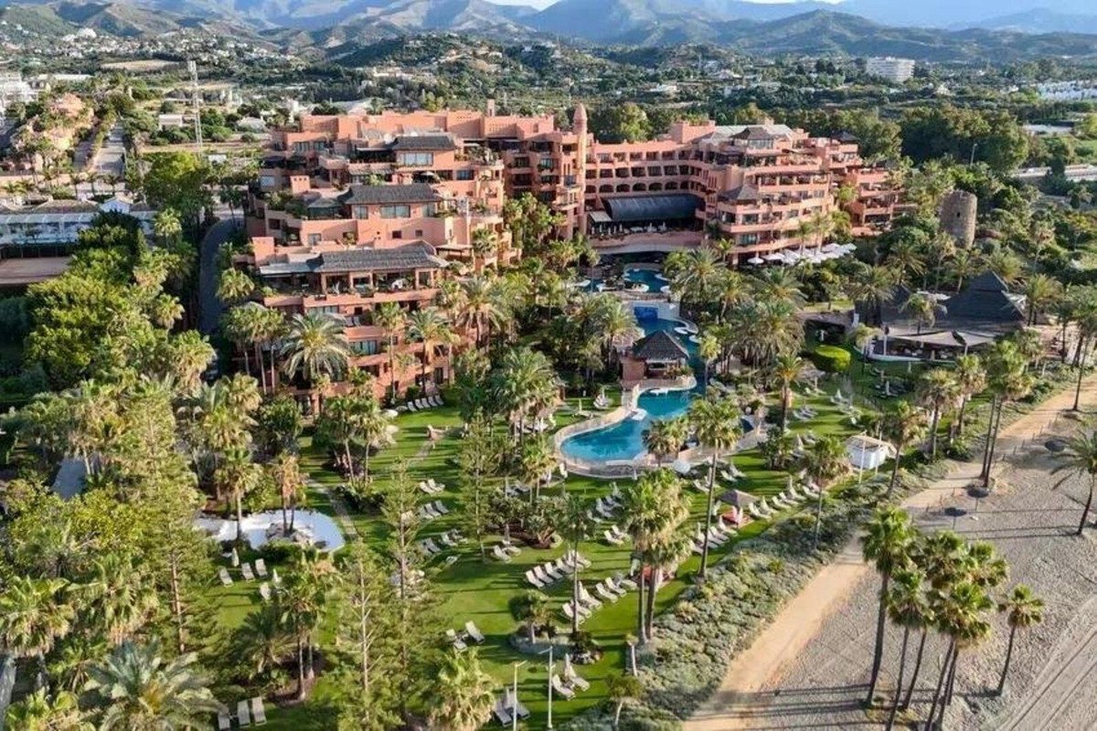 An aerial view of a resort with multiple swimming pools, lush green gardens, palm trees, and a sandy beach.