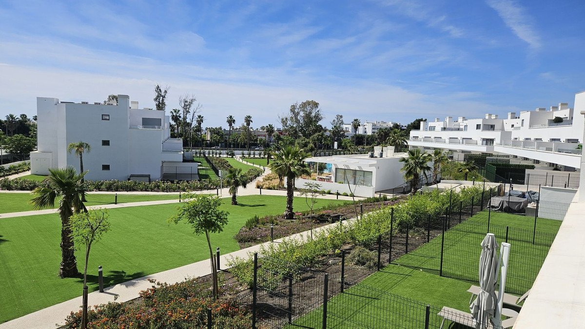 Modern white apartment complex with lush green lawns, palm trees, and a clear blue sky view.