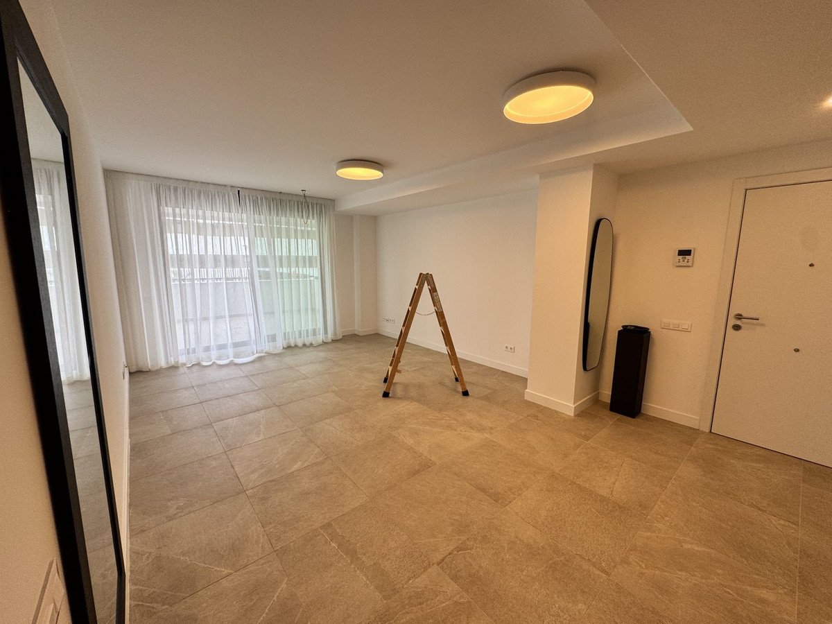 Empty living room with tiled floor, large window with sheer curtains, and ceiling lights.