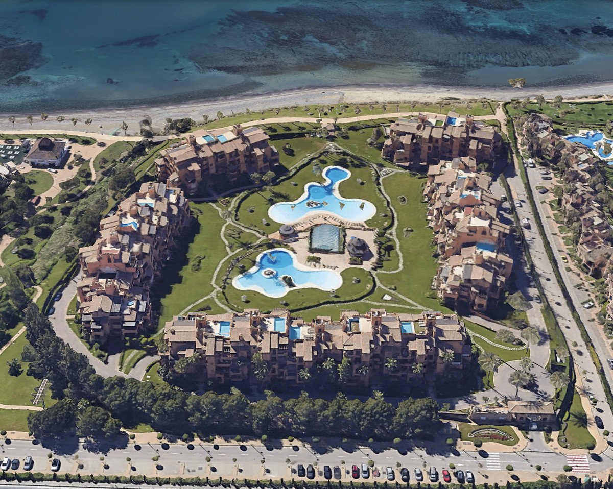 Aerial view of a coastal resort with terracotta buildings, lush green lawns, and a large kidney-shaped swimming pool near the beach.