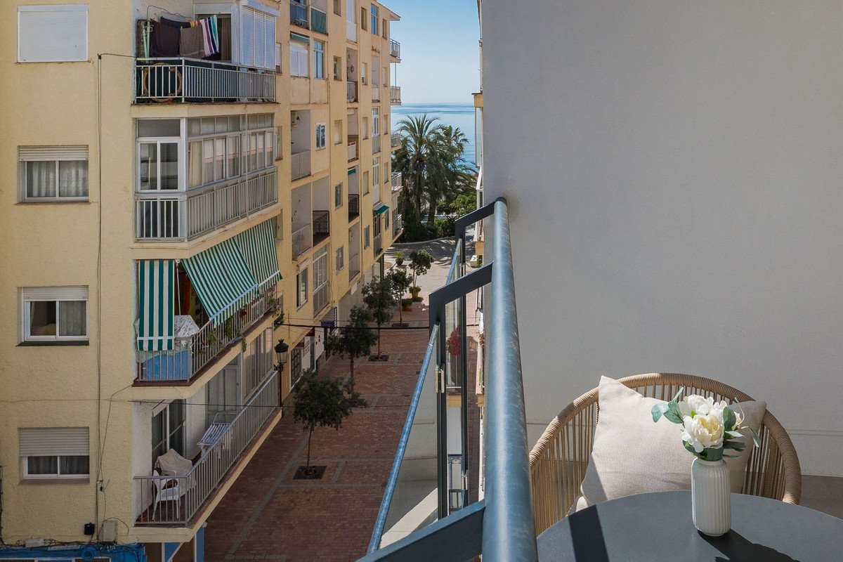 View from a balcony overlooking a street towards the sea, with palm trees and buildings. A chair and table are on the balcony.