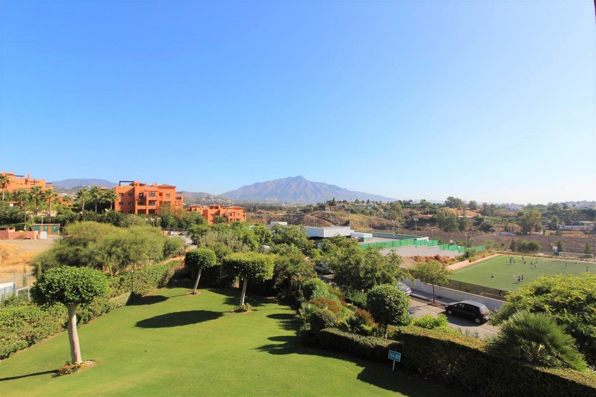 View from apartment showing a lush green garden, orange buildings, and a distant mountain under a clear blue sky.