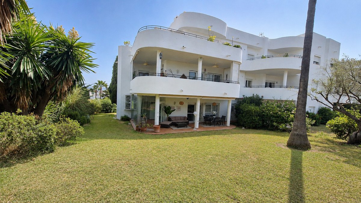 A modern white building with multiple balconies and a ground-floor terrace overlooking a lush green garden with palm trees.