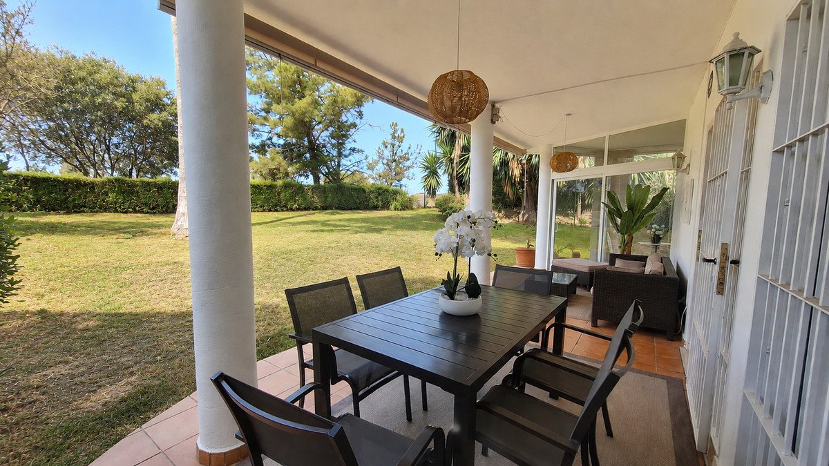 Covered patio with a dining set overlooking a lush green garden and trees under a clear blue sky.