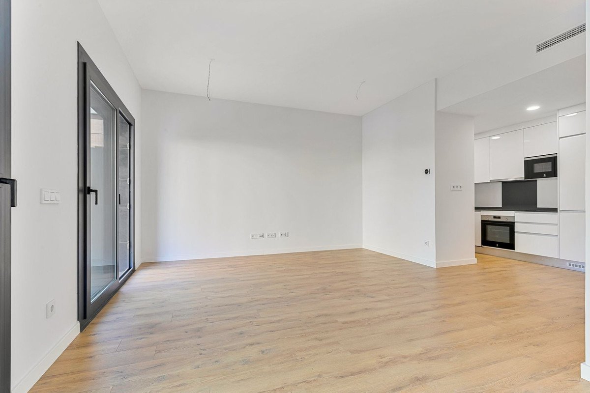 Spacious living room with light wood flooring and glass doors leading outside, open to a modern white kitchen with appliances.