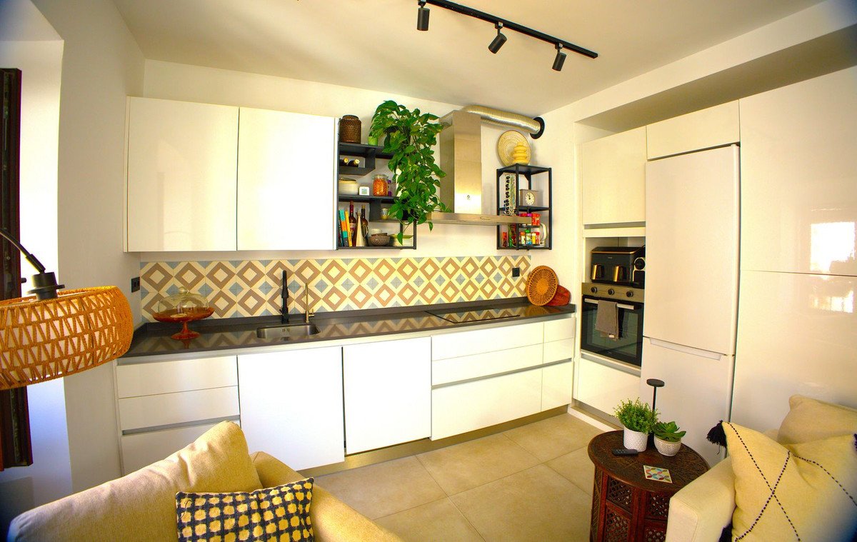 Modern white kitchen with a geometric tile backsplash, built-in oven, and refrigerator, featuring a plant and seating area.