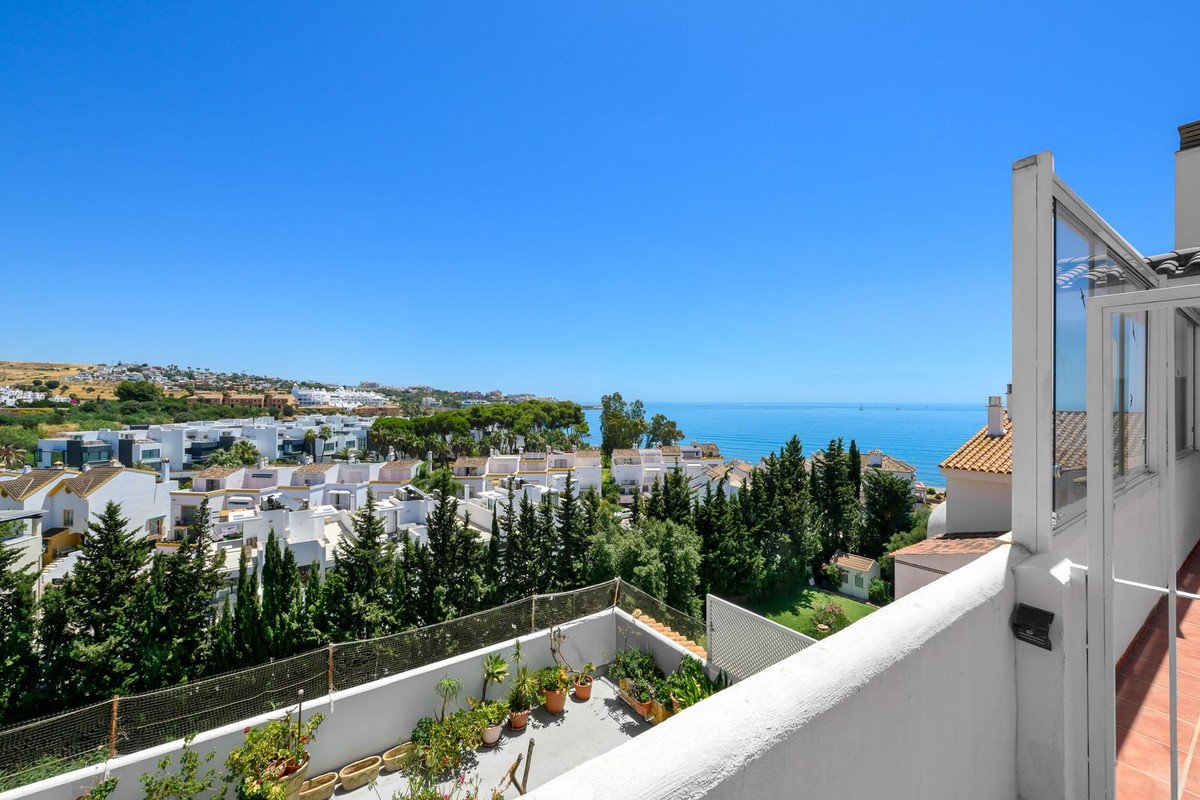 Balcony with sea view, white buildings, green trees, and clear blue sky.