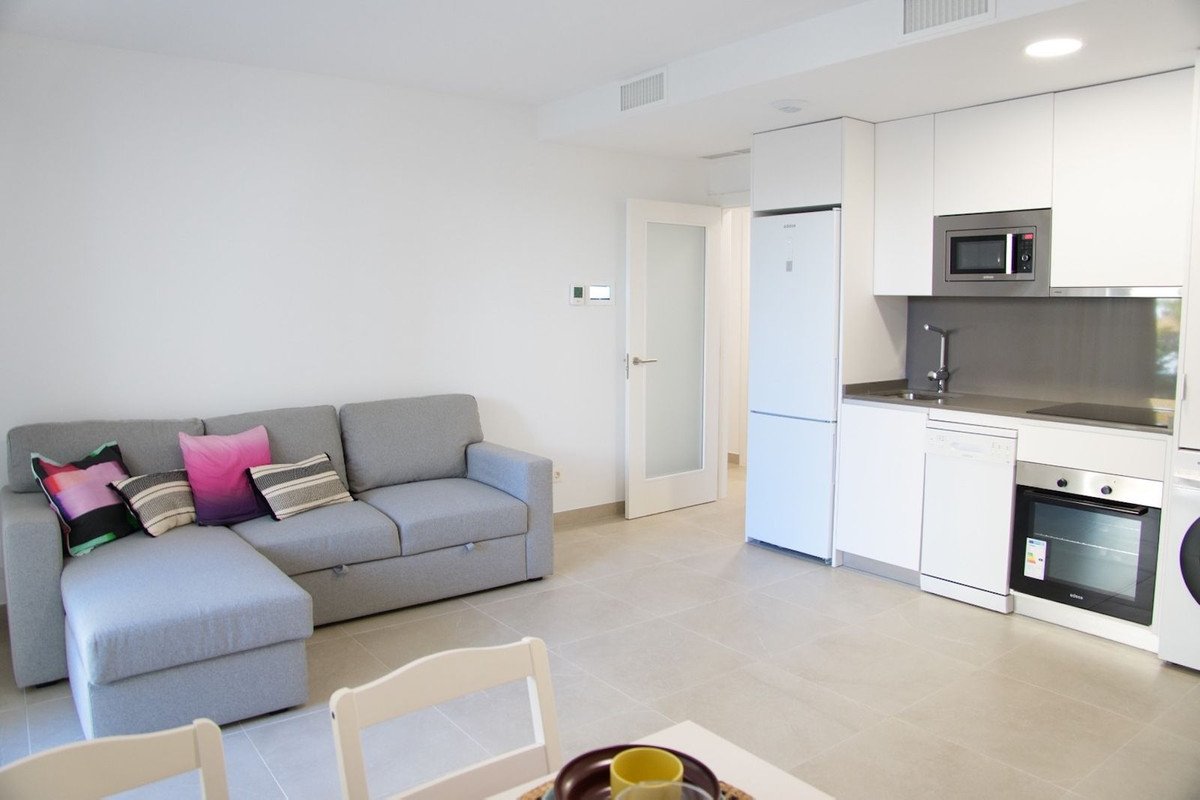A modern studio apartment living area with a grey sectional sofa and an integrated kitchen featuring white cabinets and stainless steel appliances.