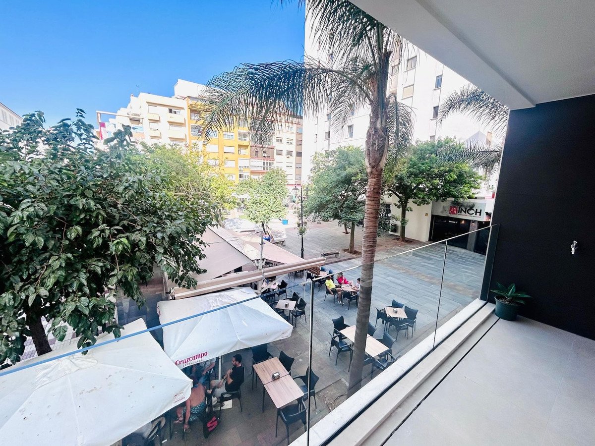 View from a balcony overlooking a street cafe with tables, umbrellas, and palm trees in a European city.