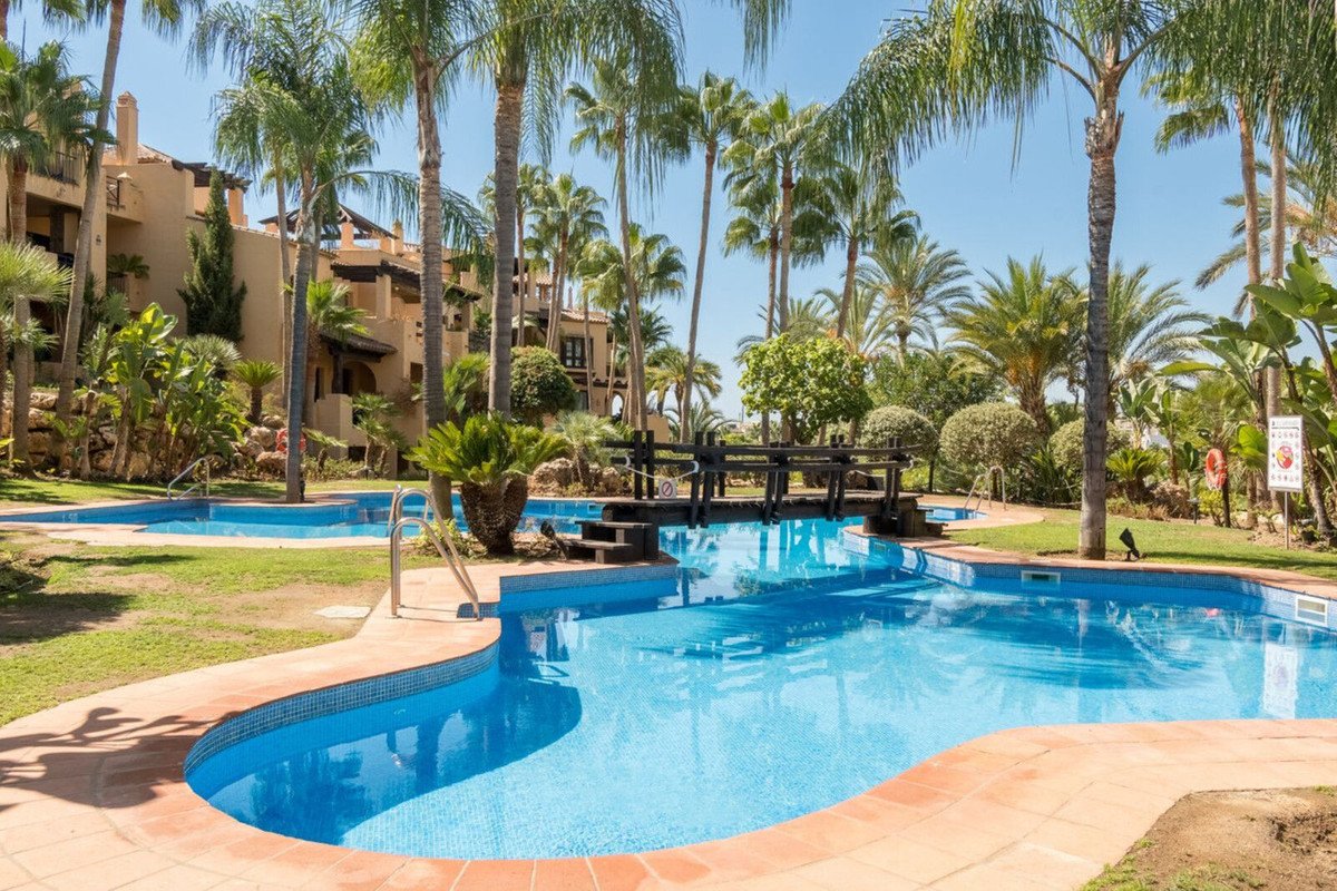 A beautiful resort swimming pool with palm trees and a decorative bridge, surrounded by lush greenery and Mediterranean-style buildings.