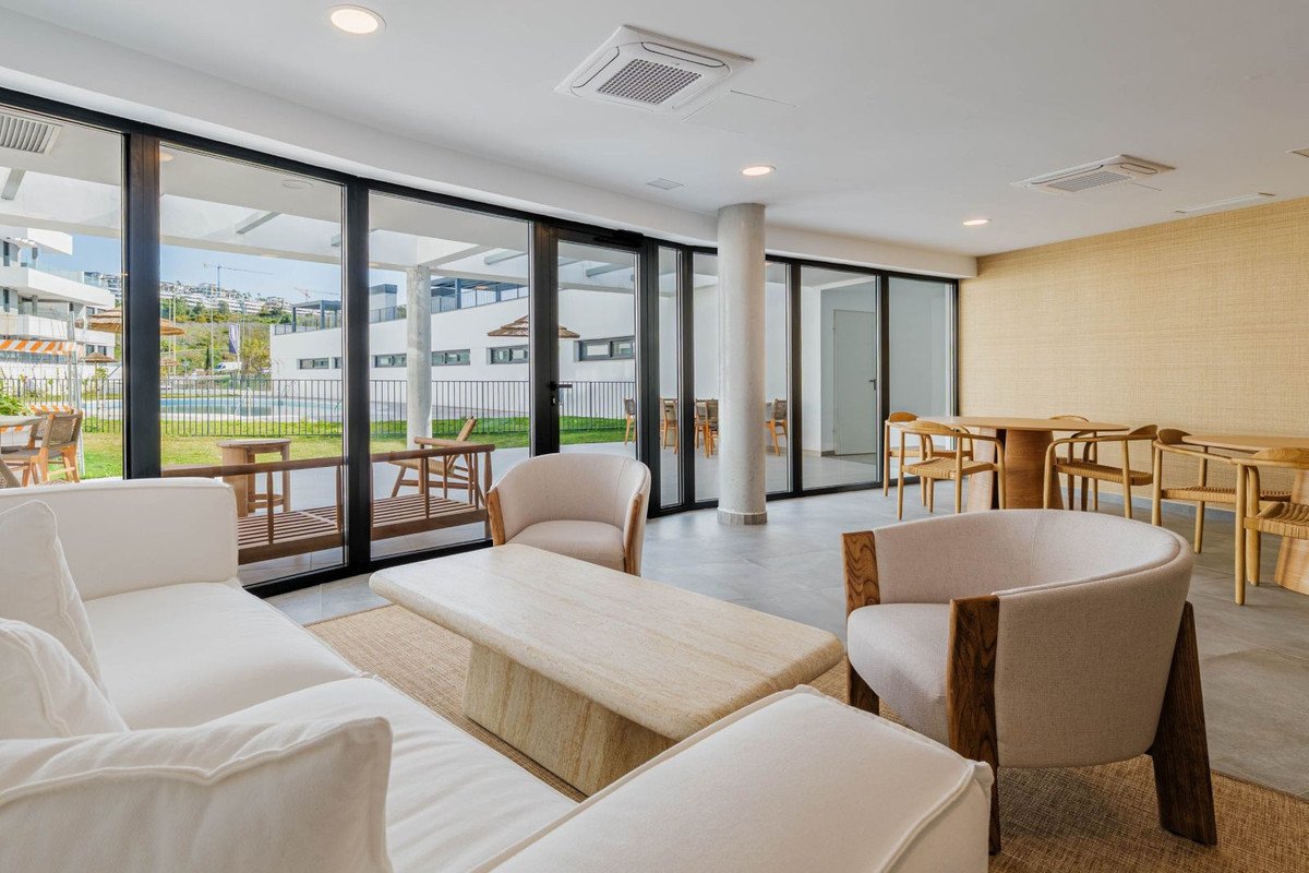 A contemporary lounge area with white sofas, chairs, and a coffee table, overlooking a swimming pool and modern buildings.