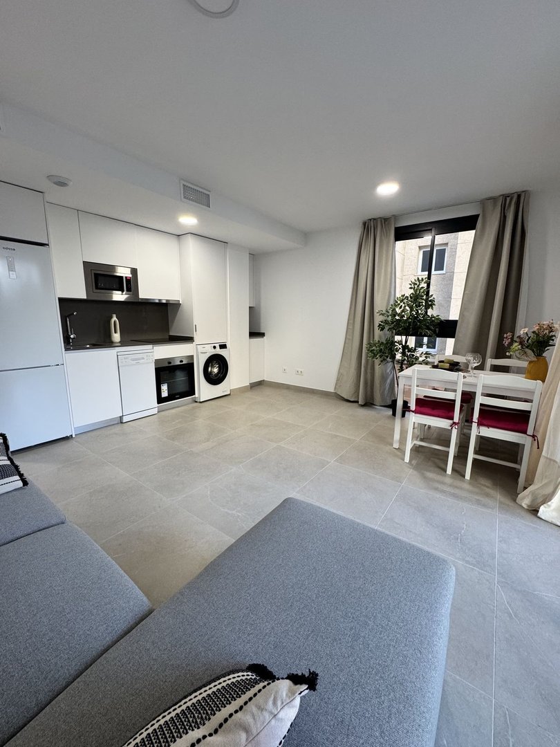 Modern studio apartment with a compact kitchenette, a dining area, and a comfortable grey sofa. A window offers city views.