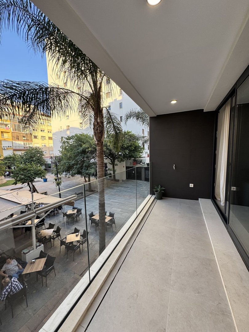 Modern balcony with glass railing overlooking a cafe patio with tables and a palm tree.
