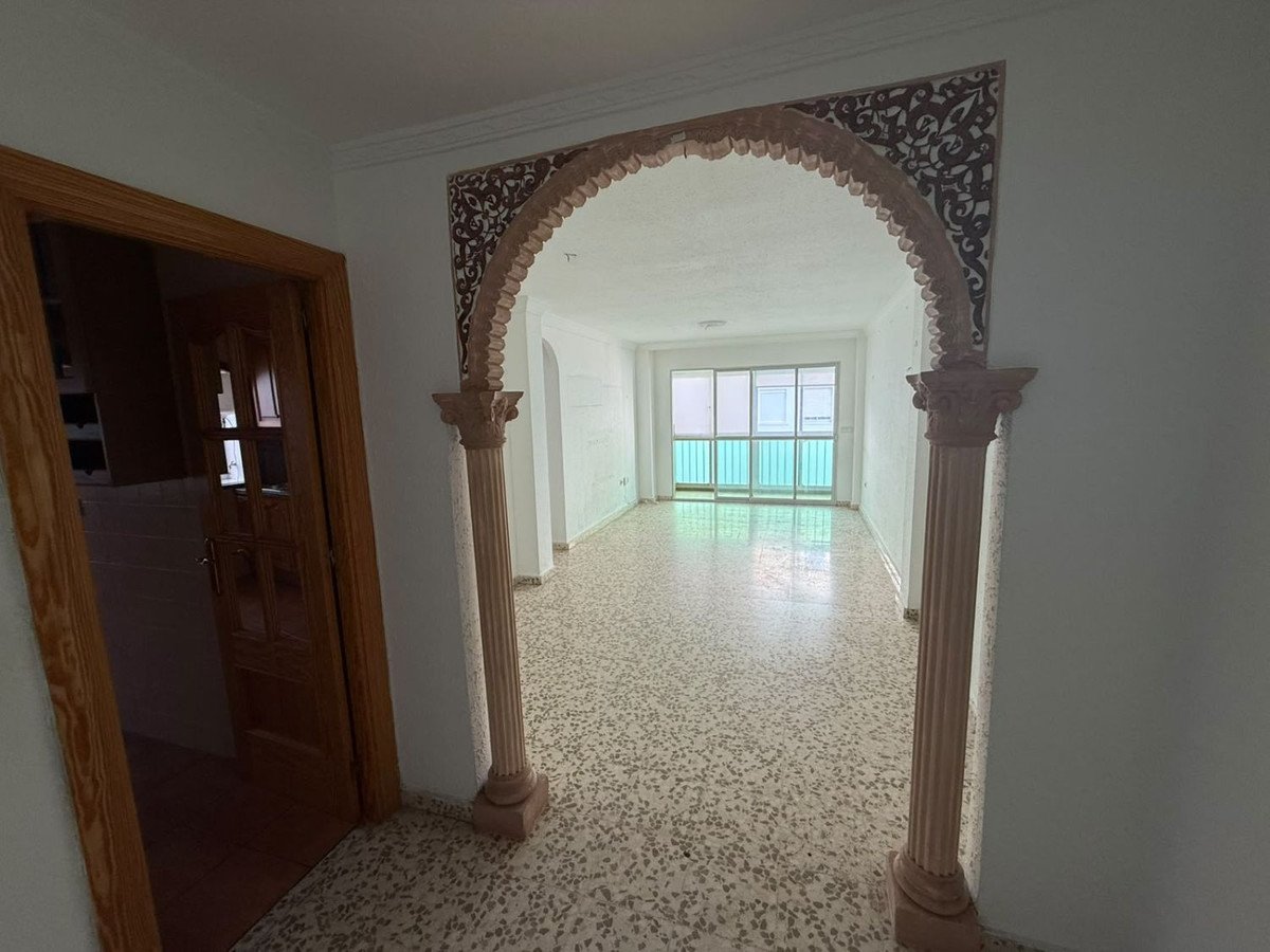 View through an ornate archway into a light-filled living room with large windows and a wooden door in the foreground.