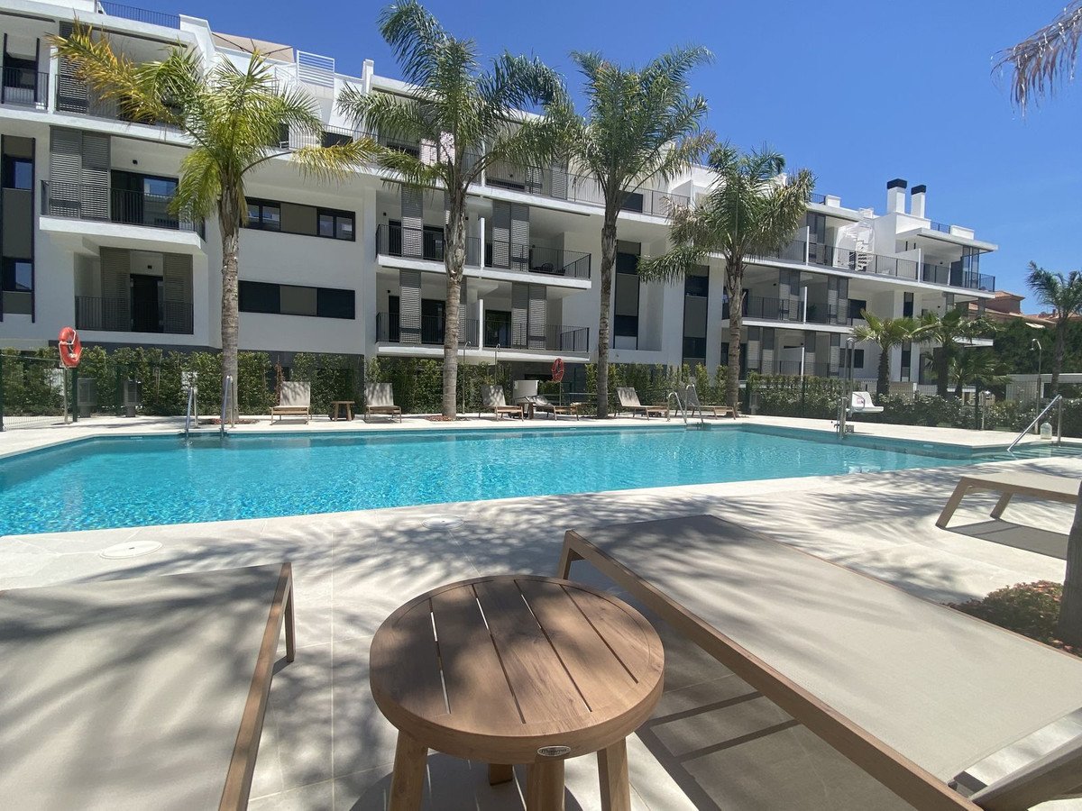 A modern apartment building overlooks a sparkling blue swimming pool surrounded by palm trees and lounge chairs on a sunny day.