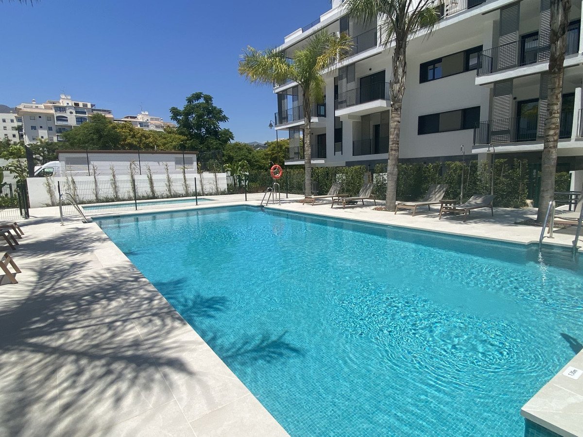 Modern apartment complex with a large swimming pool, palm trees, and lounge chairs under a clear blue sky.