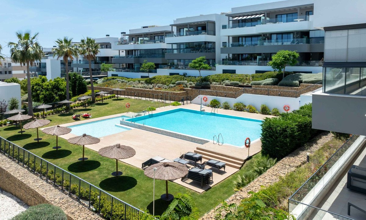 Modern apartment complex with pool, sun loungers, palm trees, and green landscaping under clear blue sky.