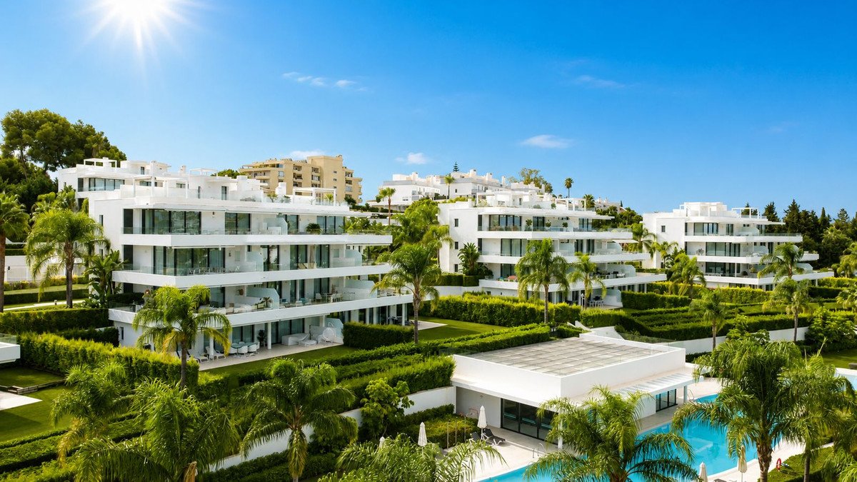Modern white apartment buildings with balconies, surrounded by lush greenery and a swimming pool under a sunny blue sky.