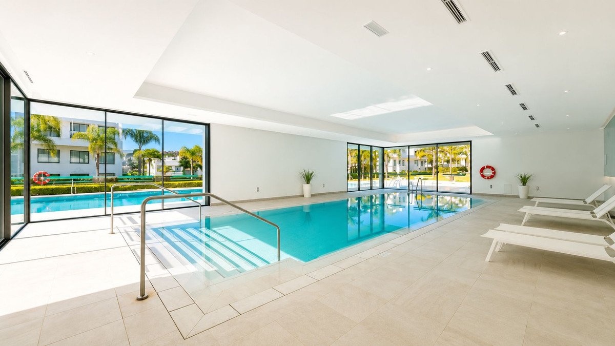 Indoor swimming pool with lounge chairs and large windows offering a view of palm trees and modern buildings.
