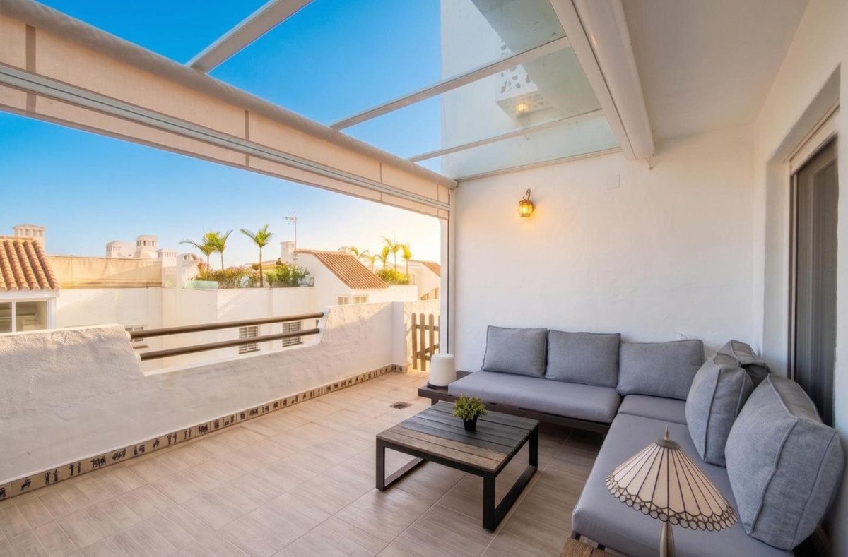 Modern rooftop terrace with gray sofa, wooden table, and panoramic view of white buildings and palm trees under a clear blue sky.