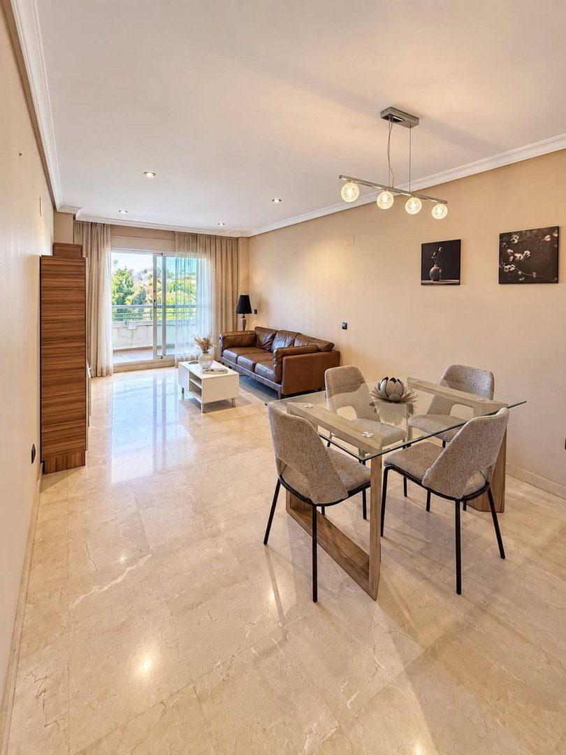 Bright living and dining area with brown leather sofa, glass dining table, and balcony access with green views.