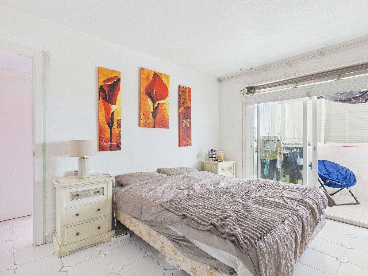 A bright bedroom with a bed, nightstands, and three floral paintings on the wall, overlooking a balcony.