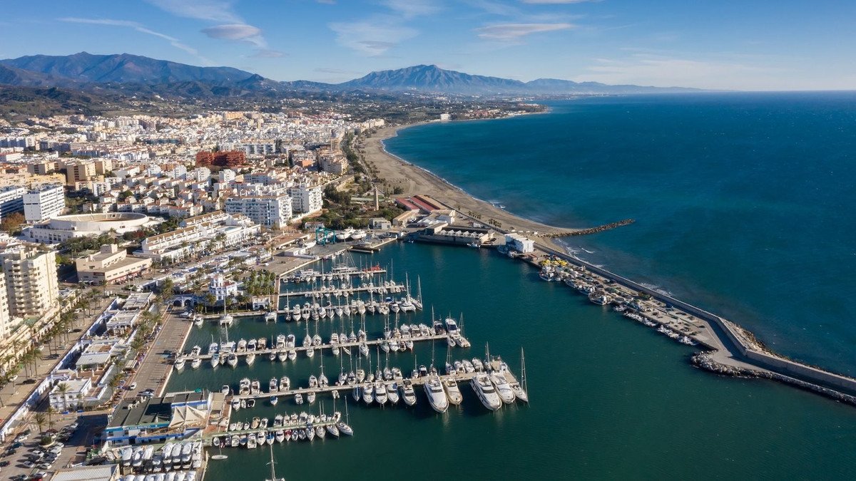 Aerial view of a coastal marina with boats, cityscape, and mountains in the background.