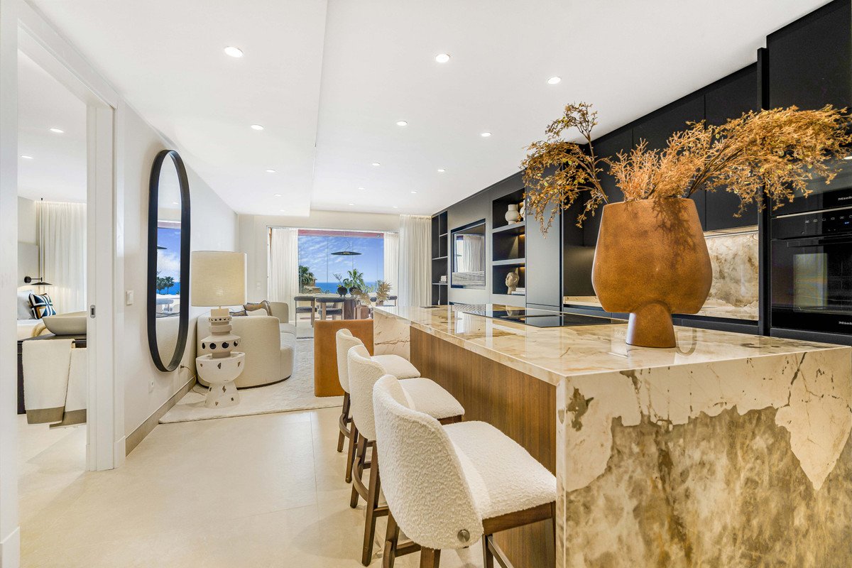 Modern kitchen island with bar stools and a living area with a large window overlooking the sea and palm trees.