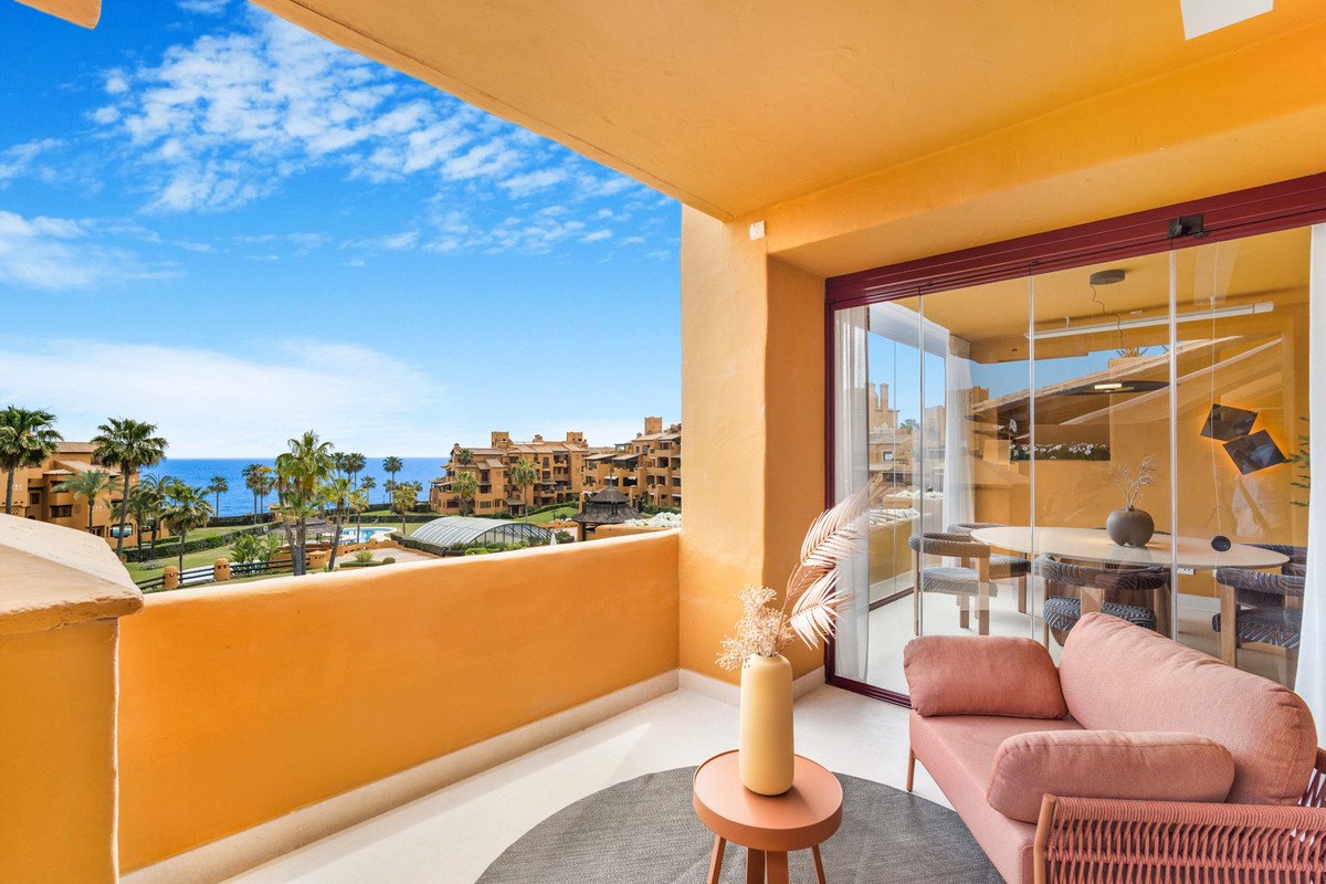 Terrace with sea view, sofa, and dining area visible through glass doors. Palm trees and resort buildings in the background.