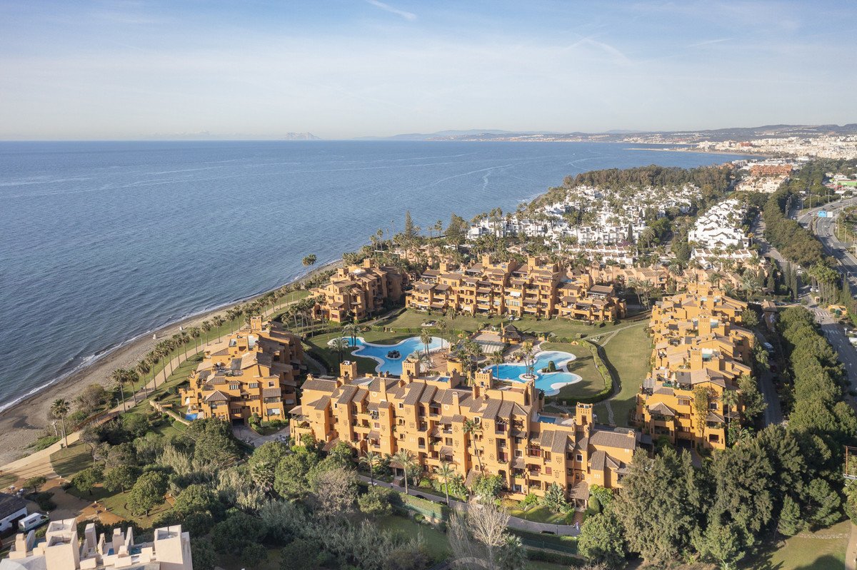 Aerial view of an apartment complex with multiple buildings, swimming pools, green lawns, and the sea in the background.
