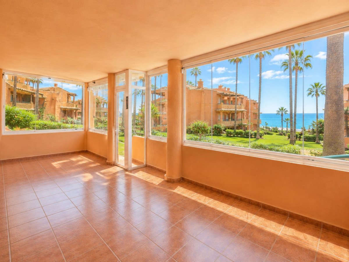 Enclosed balcony with floor-to-ceiling windows offering stunning sea views, palm trees, and resort buildings.