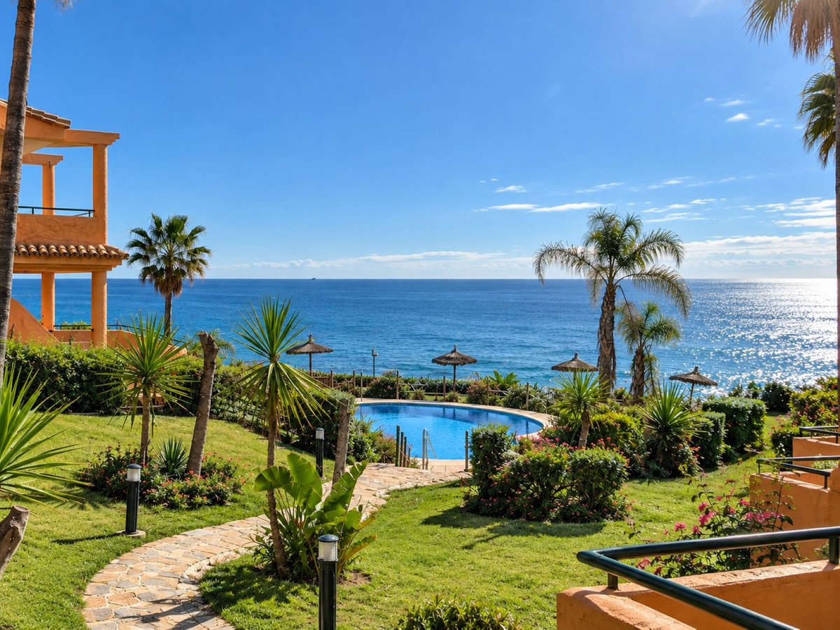 Resort swimming pool with loungers and umbrellas, surrounded by tropical gardens and palm trees with the blue sea in the background.