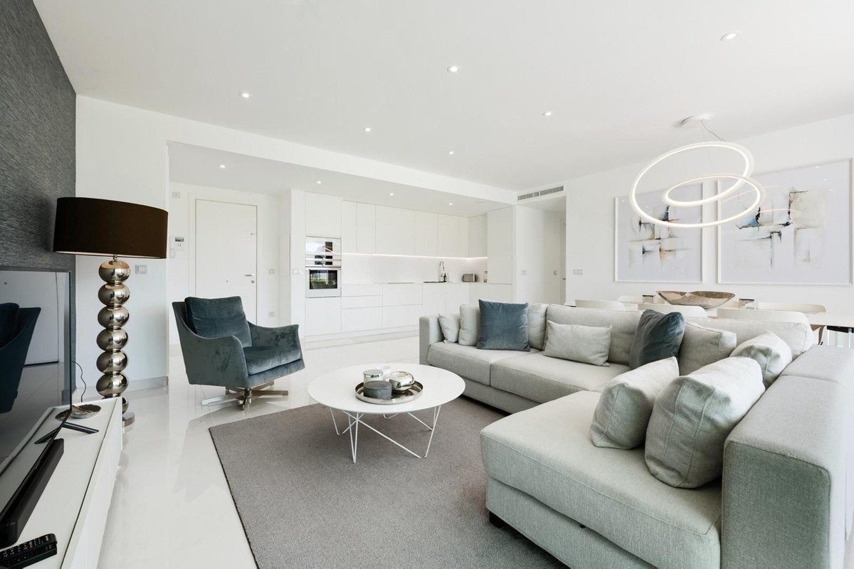 A spacious modern living room featuring a large sectional sofa, armchair, coffee table, and an open-plan kitchenette with white cabinetry and dining area.
