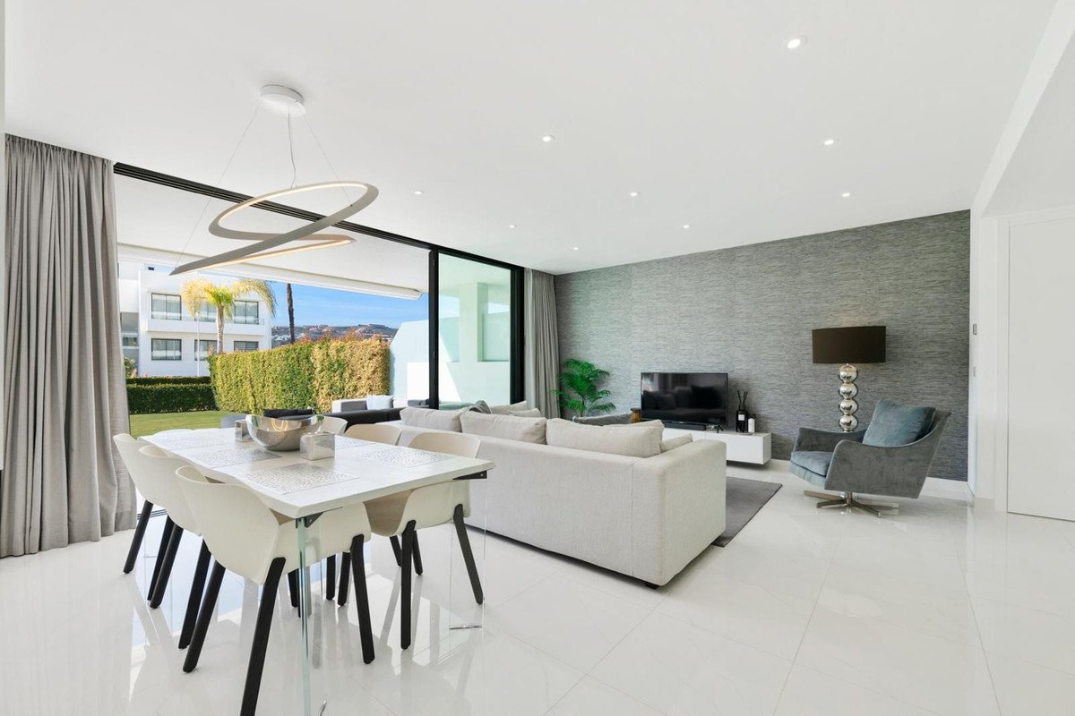 Modern living and dining area with a glass table, white sofa, and a view of the garden and hills.