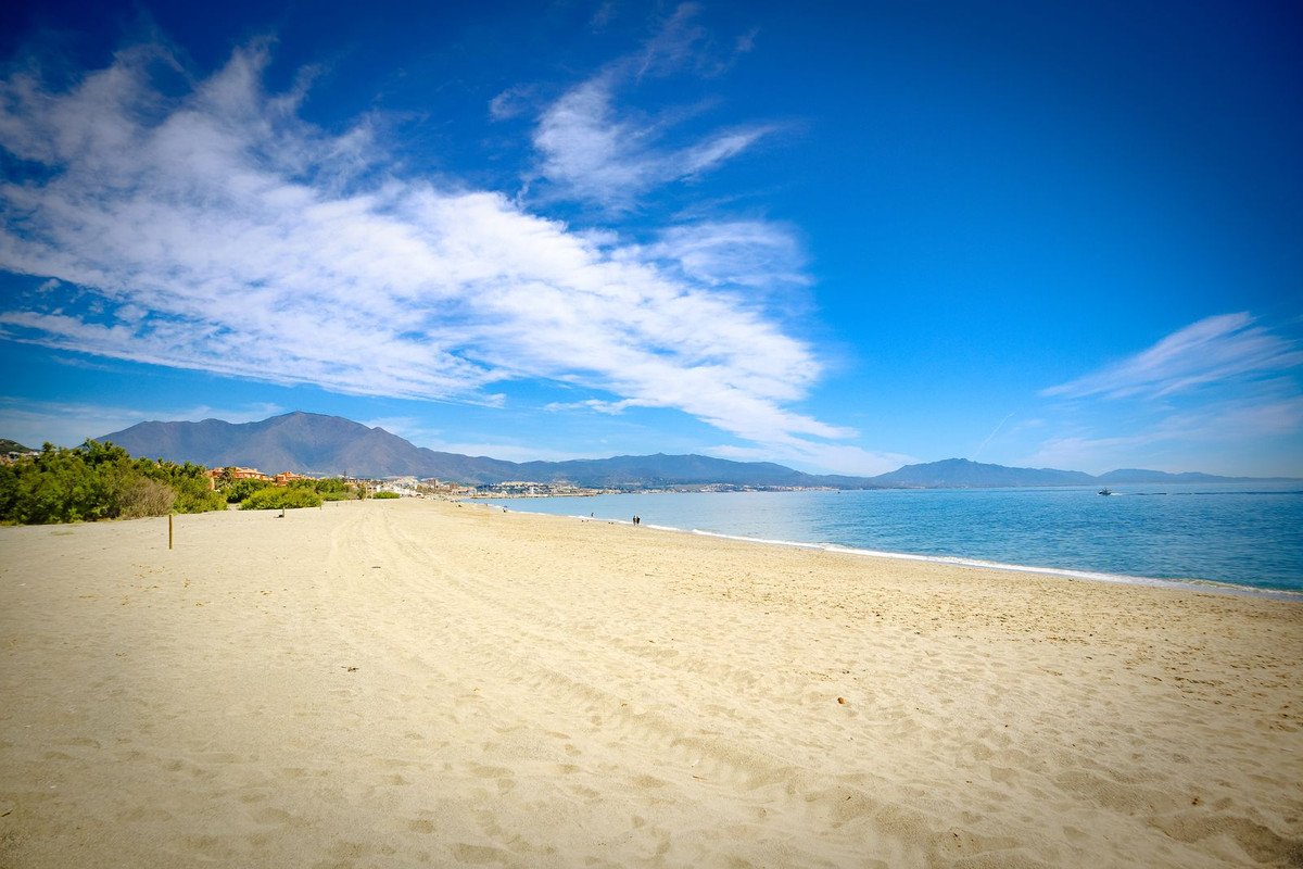 Expansive sandy beach with calm blue sea and distant mountains under a bright blue sky with white clouds.