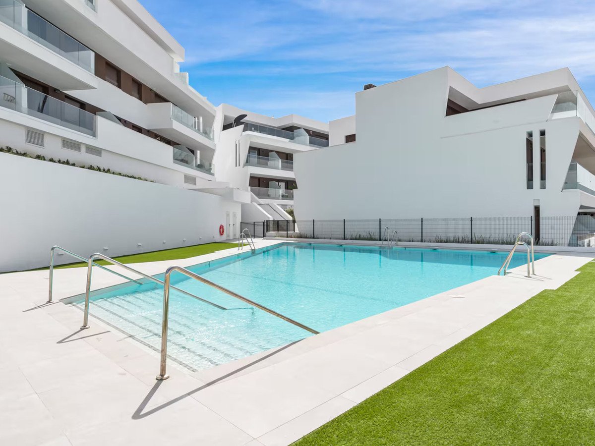 A large, modern swimming pool with stairs and handrails, surrounded by white buildings and green artificial turf.