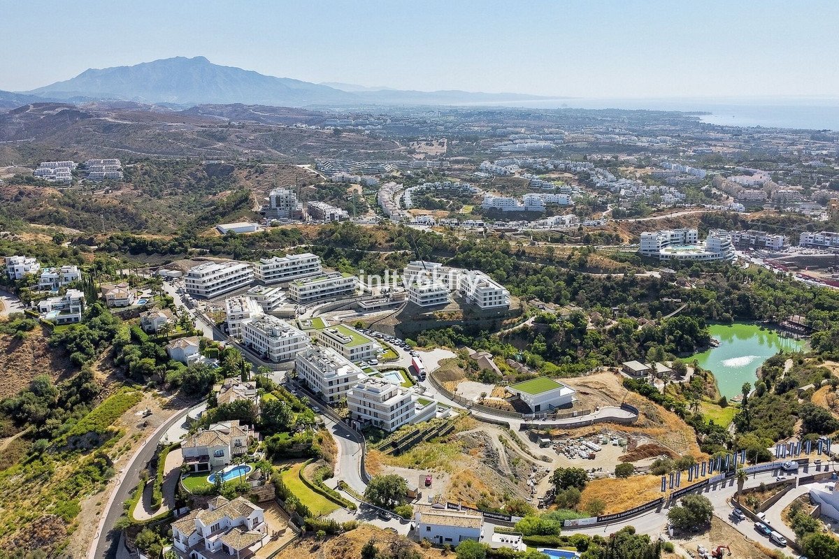 Aerial view of modern apartment buildings with swimming pools and a lake, overlooking a distant sea and mountains.