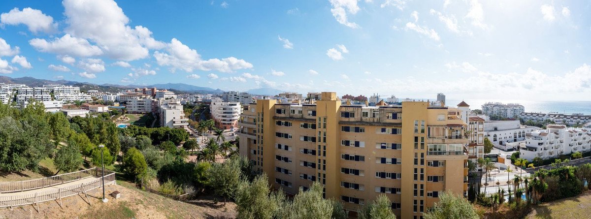 Panoramic view of apartment buildings with trees and the sea in the background under a blue sky with clouds.