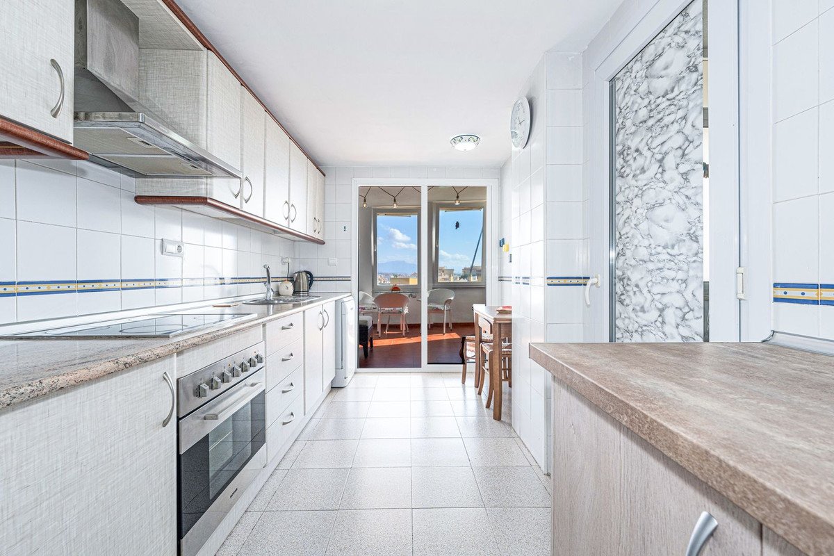 A long galley kitchen with white cabinets and tiled walls, opening to a balcony with a table and a table and chairs and a distant view.