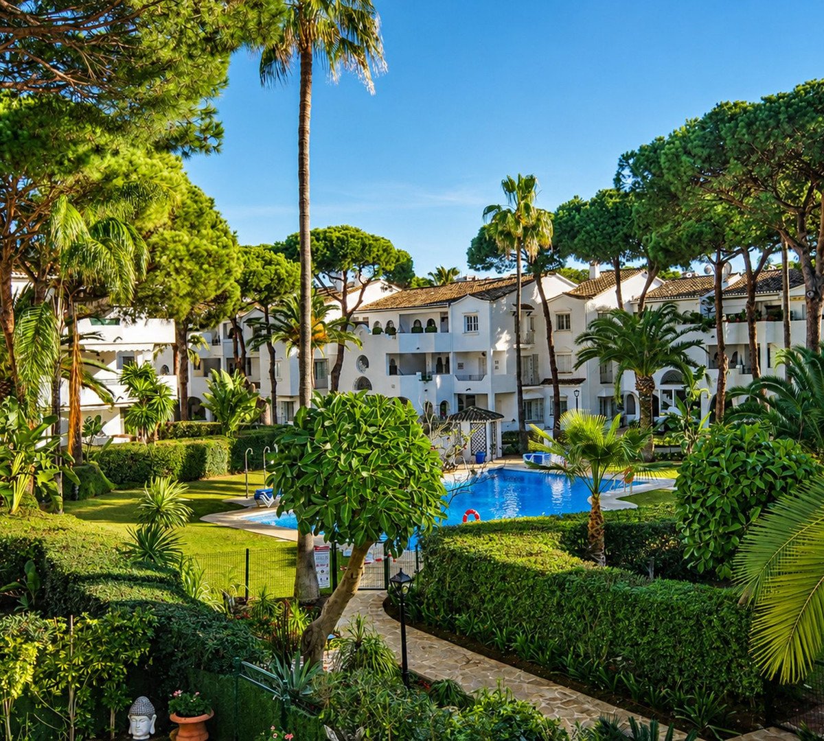 Villa with pool view, lush greenery, palm trees, and white buildings under a clear blue sky.
