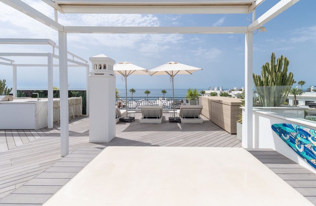 Rooftop terrace with lounge chairs, umbrellas, and ocean view. White pergola, wooden deck, and cactus accents.